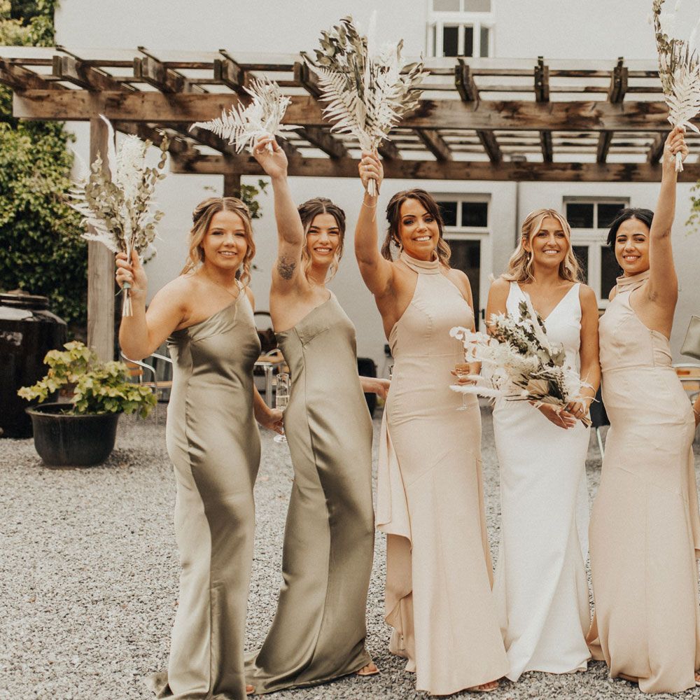 Bride in white V neck Made With Love wedding dress holding white and green dried bridal bouquet stands with bridesmaids in satin sage green and high necked beige bridesmaids dresses whilst they wave fern bridesmaids bouquet at Anran Devon