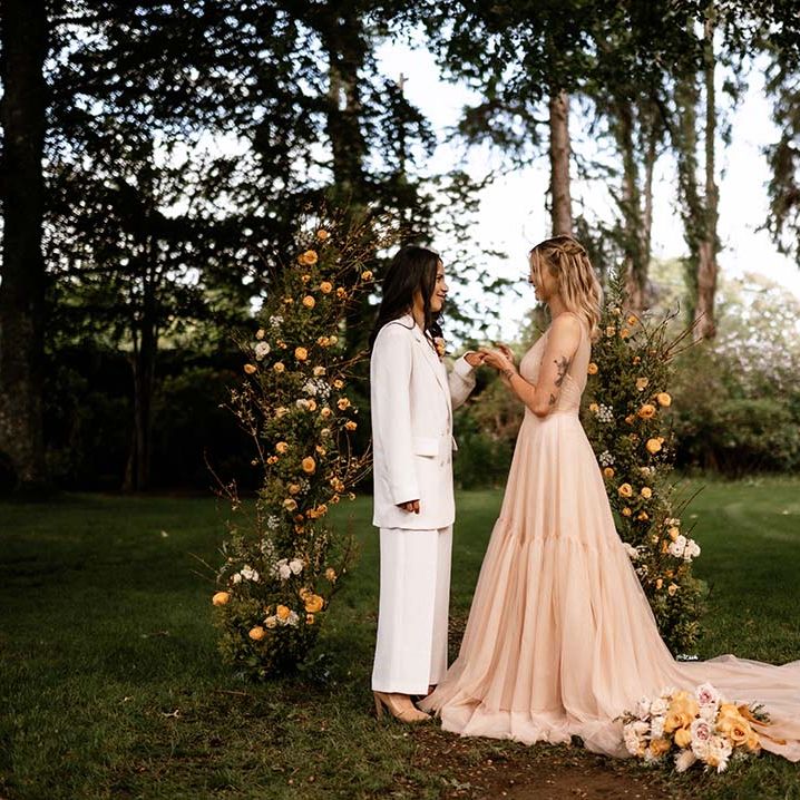 Outdoor wedding ceremony with two brides in a white suit and blush pink dress exchanging vows 