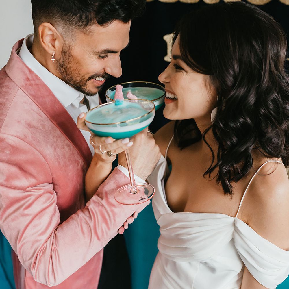 Groom in a pink jacket and bride in a cold shoulder wedding dress drinking blue cocktails with sweets in 