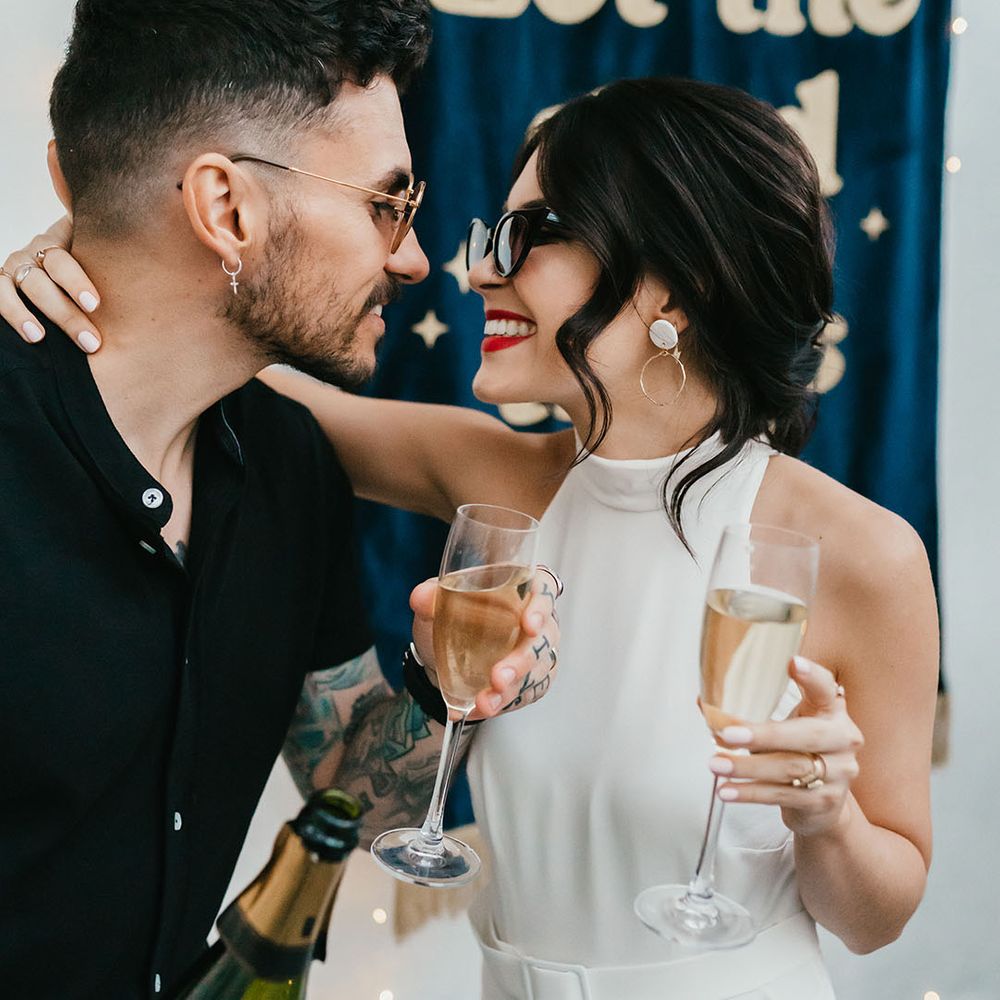 Bride with pinned up hair wearing sunglasses, hoop earrings and red lipstick smiling at her groom in a black shirt whilst they sip champagne 