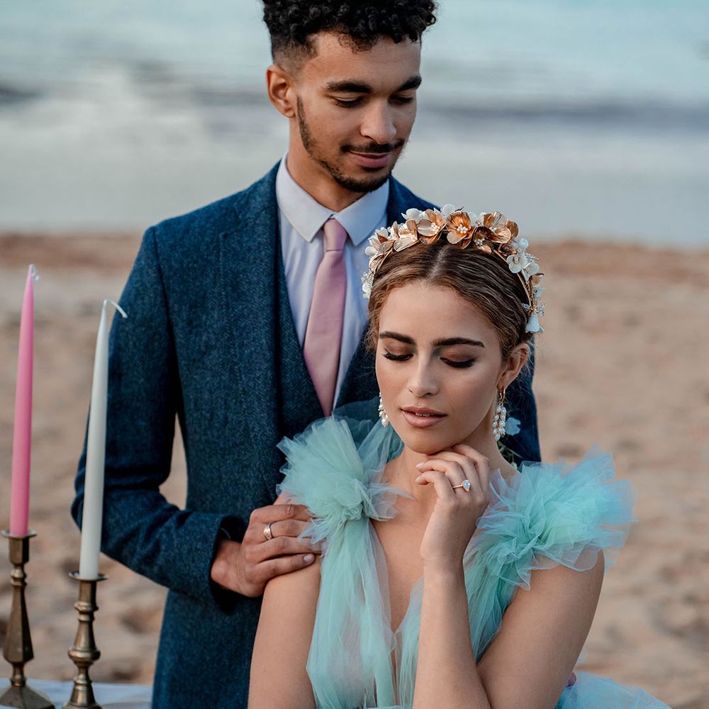Bride in a mint green wedding dress sitting at a relaxed tabelscape on the beach with her groom in a navy blue suit and pink tie standing behind her