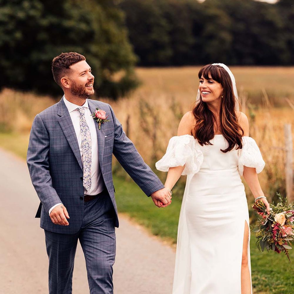 Groom in a blue check suit holding hands with his bride in a strapless wedding dress with lace puff sleeves and front split design 