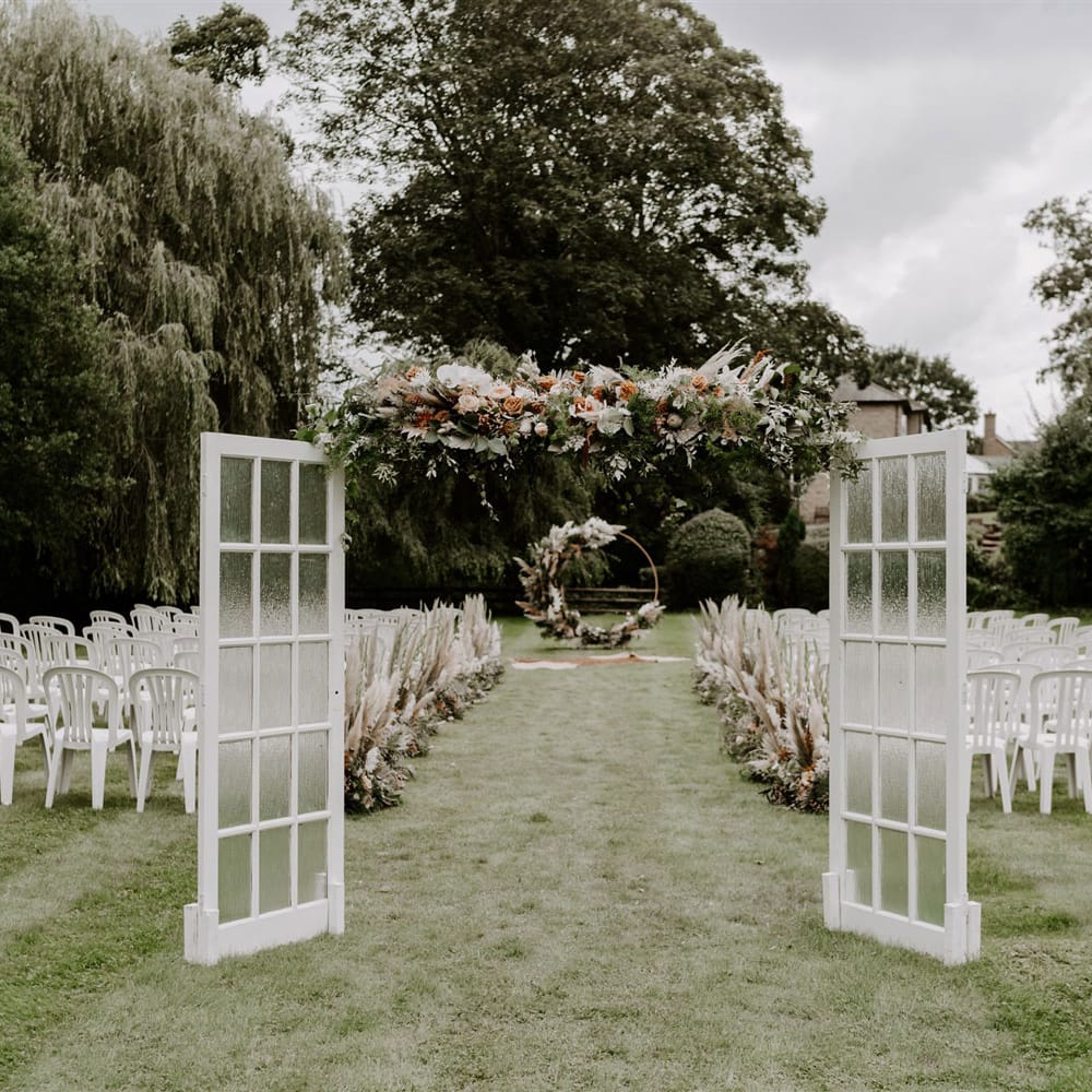 Green glass doors and floral garland open onto garden wedding ceremony with white chairs, pampas grass floral installations and floral Moorgate for DIY garden wedding