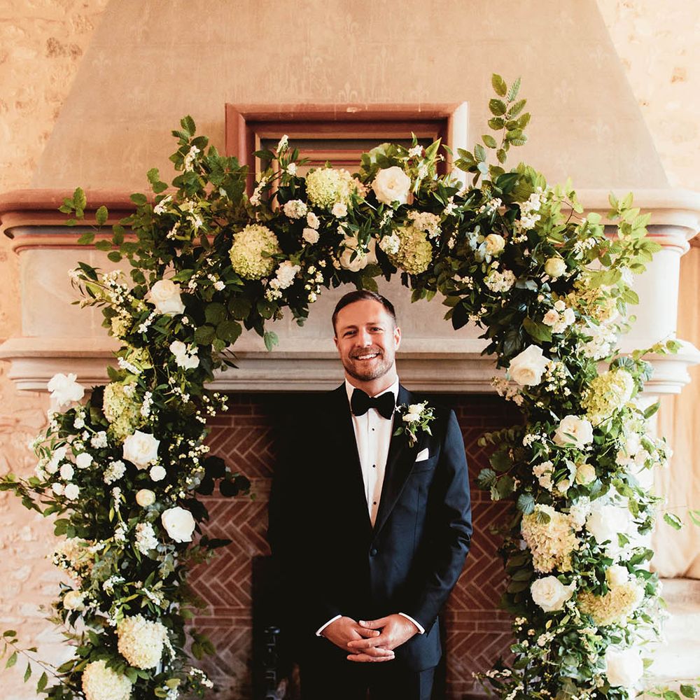 Groom stands in front of floral archway 