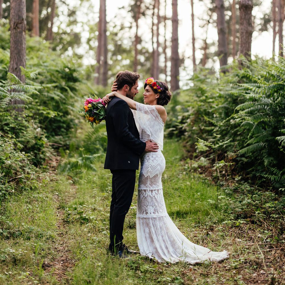 Bride in off the shoulder Grace Loves Lace wedding dress and flower crown holding colourful wedding bouquet hung groom in ball suit in woodland after outdoor wedding ceremony