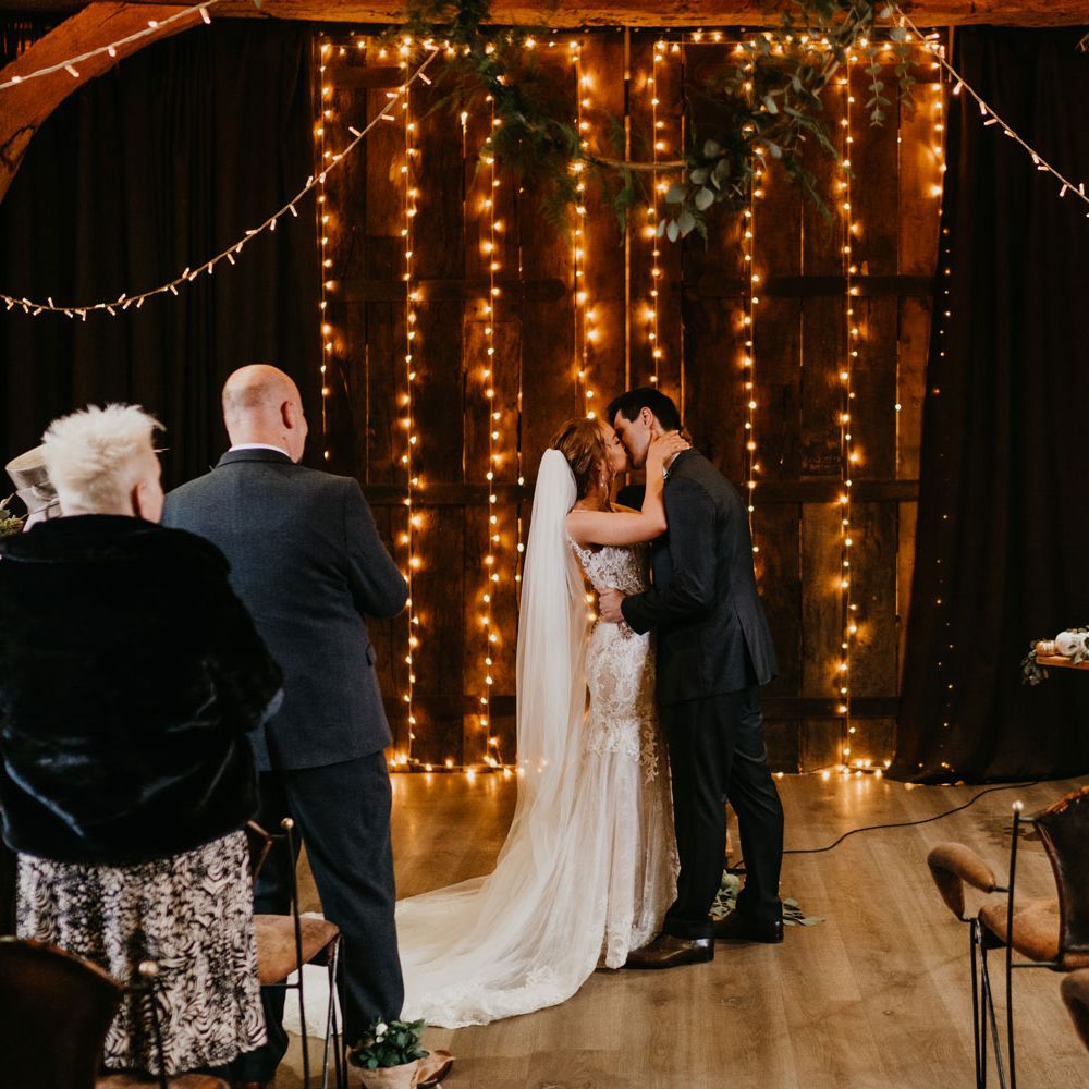 Bride in tulle wedding dress with train and veil kisses groom in dark suit in front of fairy lights for barn wedding ceremony