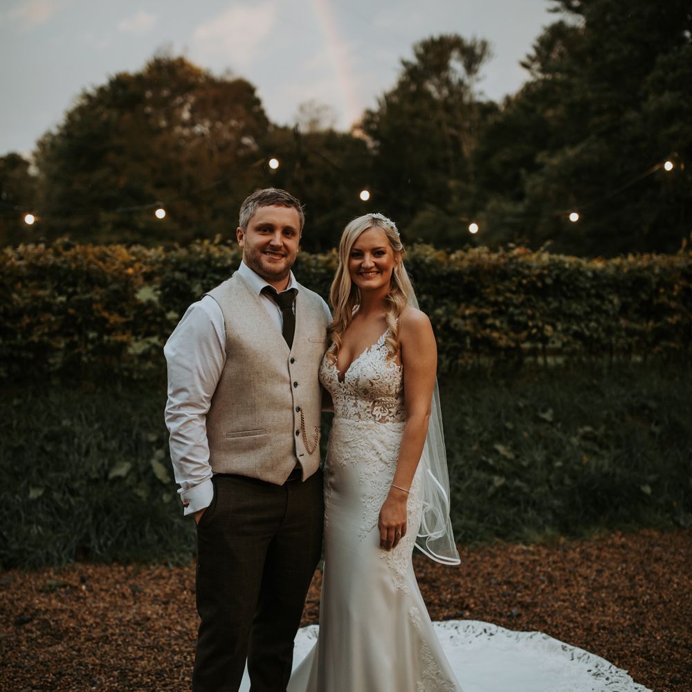 Bride in lace wedding dress with train and veil stands with groom in beige waistcoat outside with rainbow in the sky