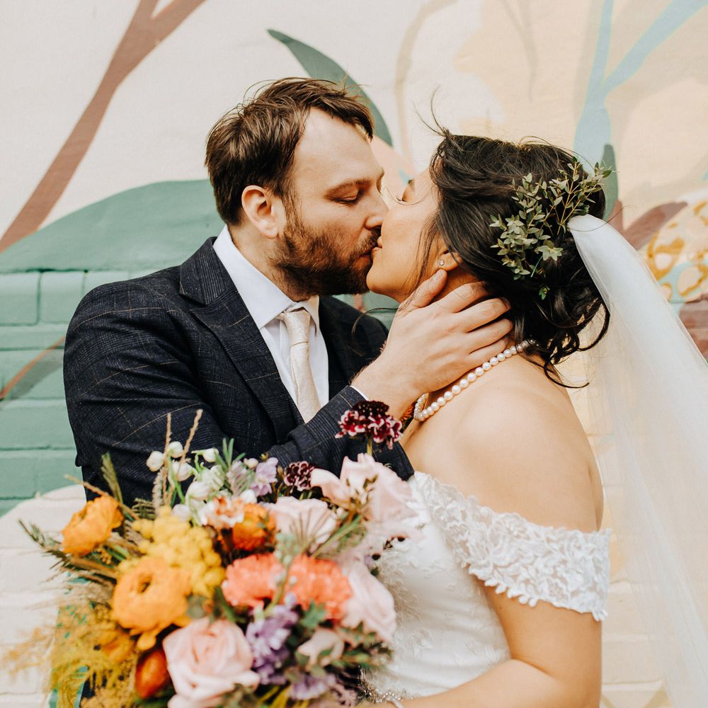 Groom in blue check suit with light pink tie kisses bride in off the shoulder lace dress and pearl necklace holding orange, yellow and pink bouquet by street art at Scale in Liverpool