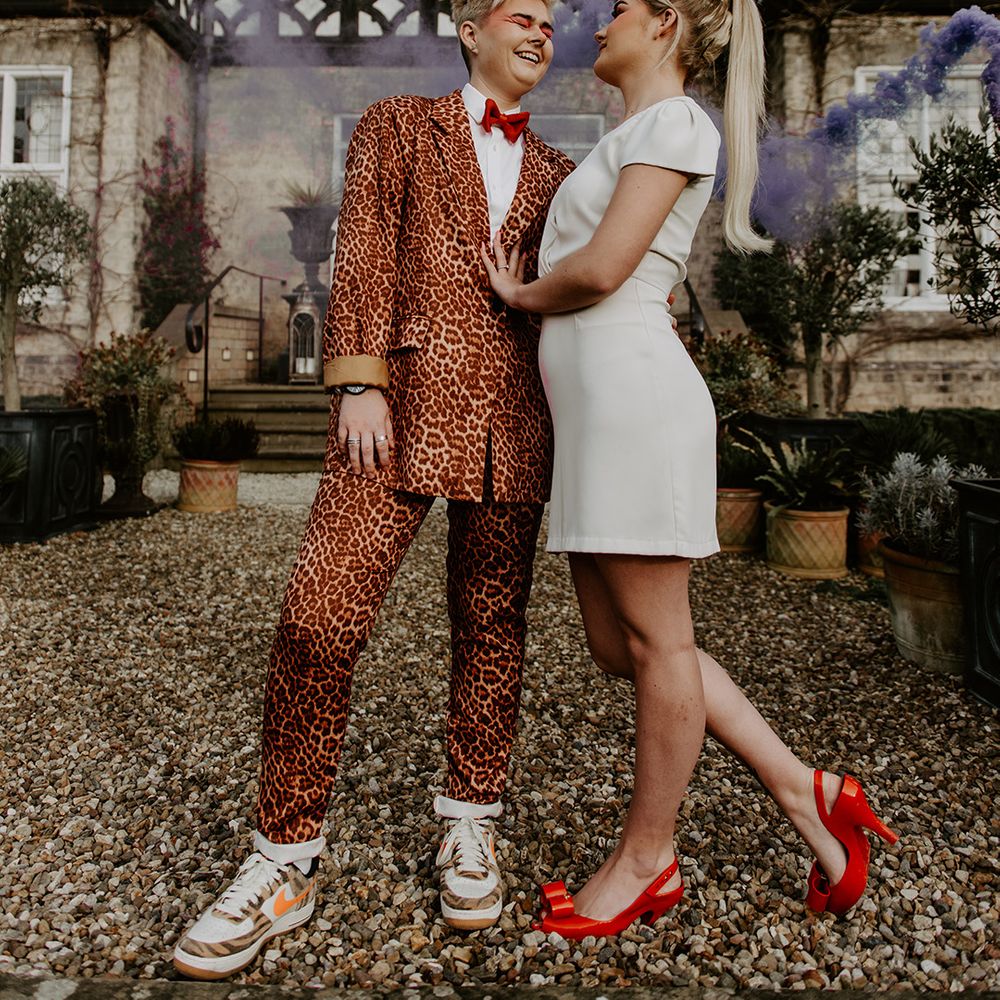 LGBTQ+ wedding with bride in a leopard print suit and trainers and a short wedding dress with bow in her hair with a purple smoke bomb going off in the background