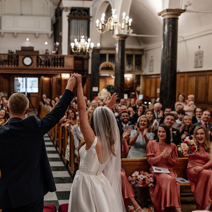 Bride in a princess wedding dress with long veil and groom in a navy suit holding their hands in the air at the church altar 