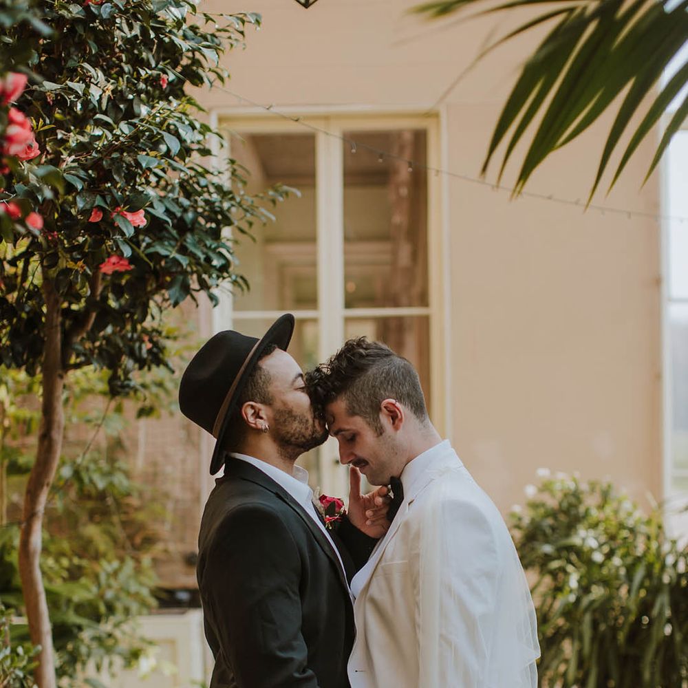 Groom in a black suit and fedora hat kissing his partner head in a white groom suit and cape