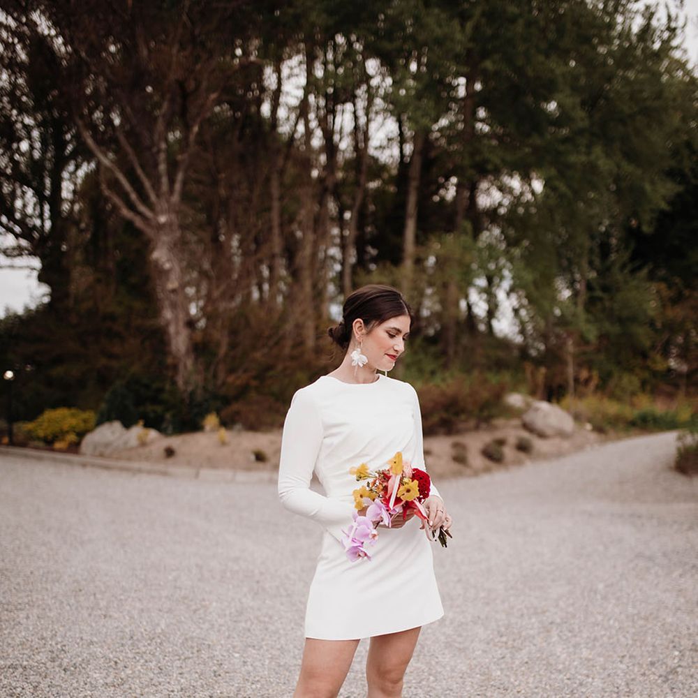 Bride in a short wedding dress with long sleeves and fuchsia pink wedding shoes holding a red, pink and yellow bouquet with orchids
