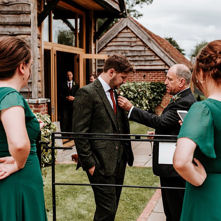 Bridesmaids in emerald green dresses stand with the groom and father as he fixes the groom's buttonhole 