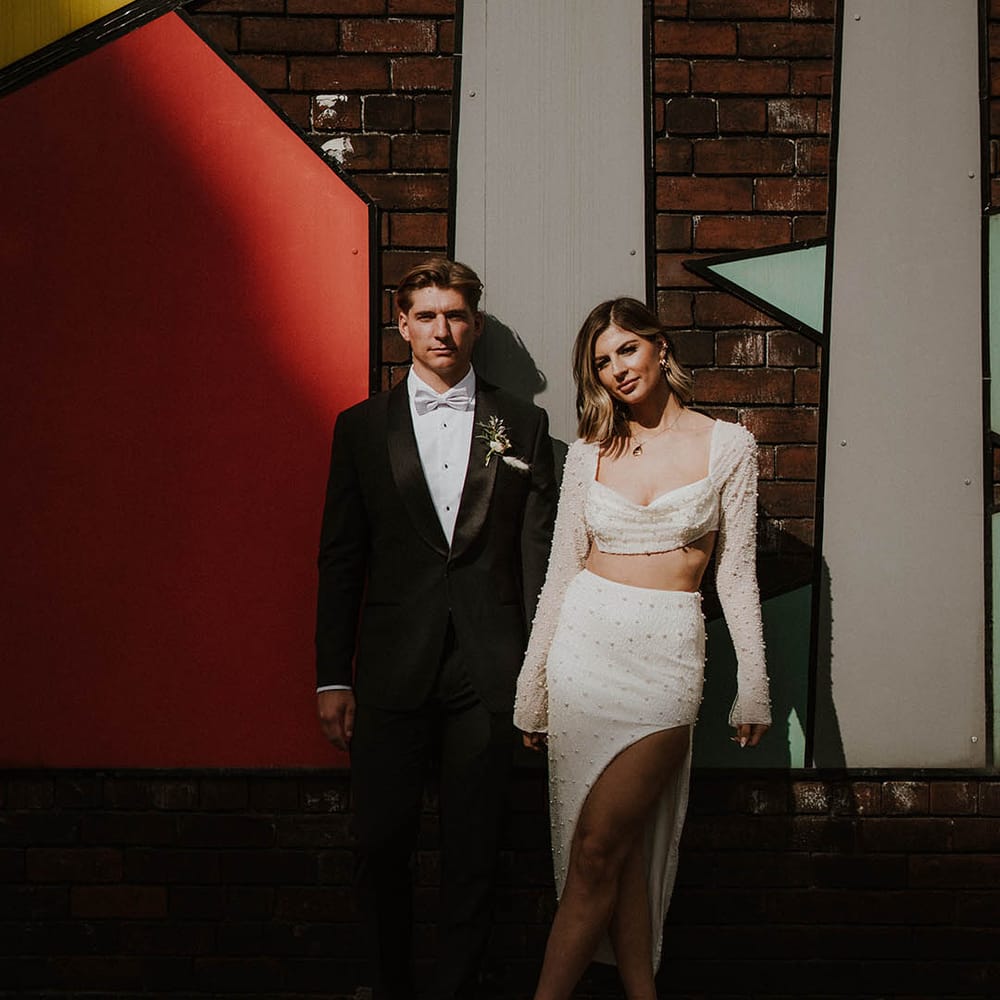 Bride and groom pose together with groom in suit jacket with satin lapels and pale blue bow tie and bride in two piece pearl outfit and white feather shoes