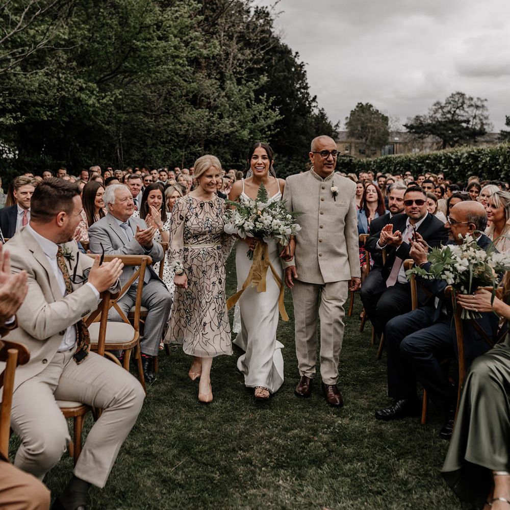 Mother and father of the bride walking their daughter down the aisle at her outdoor back garden wedding 