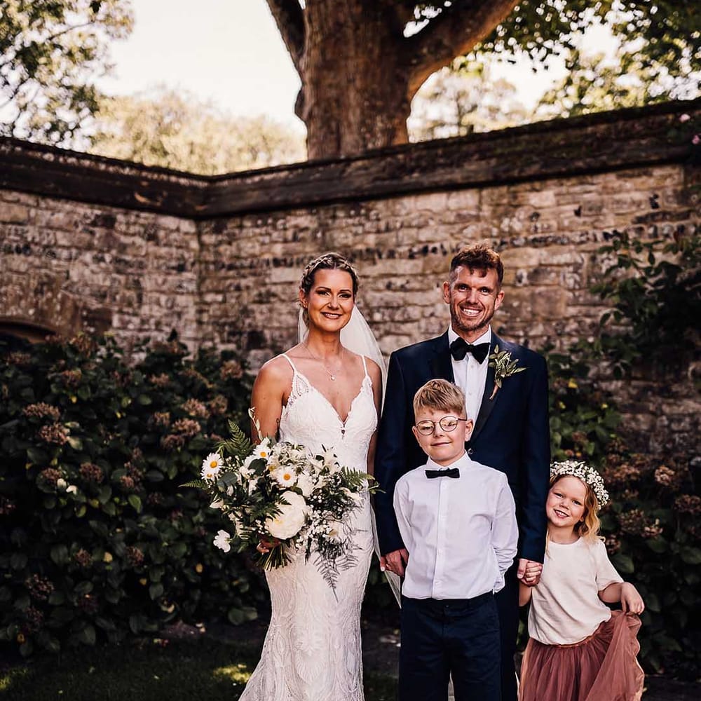 Bride and groom stand holding hands with their son and daughter for outdoor wedding ceremony 