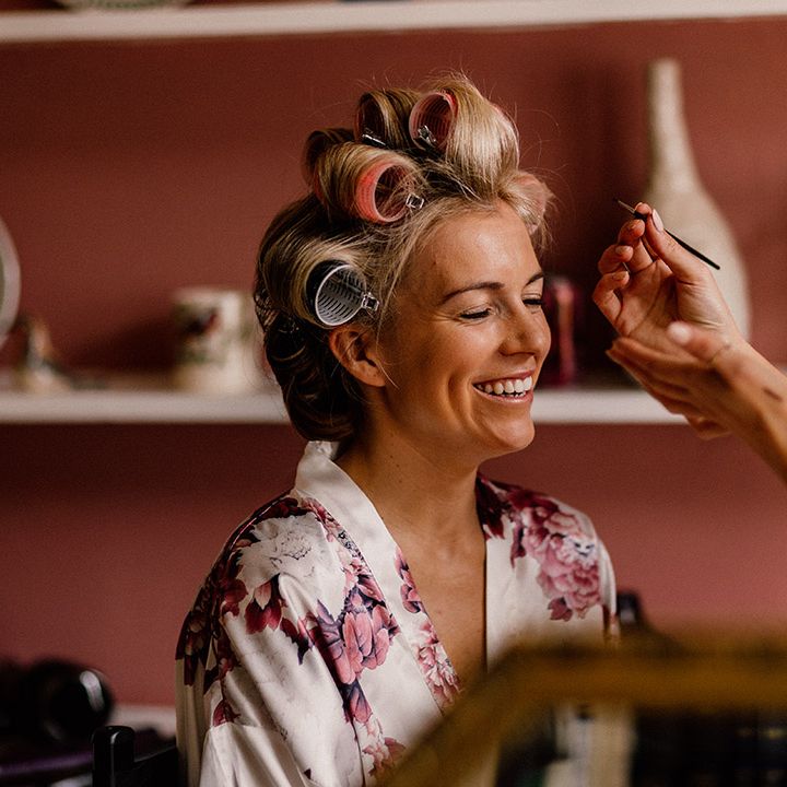 Bride in pink and white floral satin robe gets her makeup done for the wedding day with her hair in rollers 