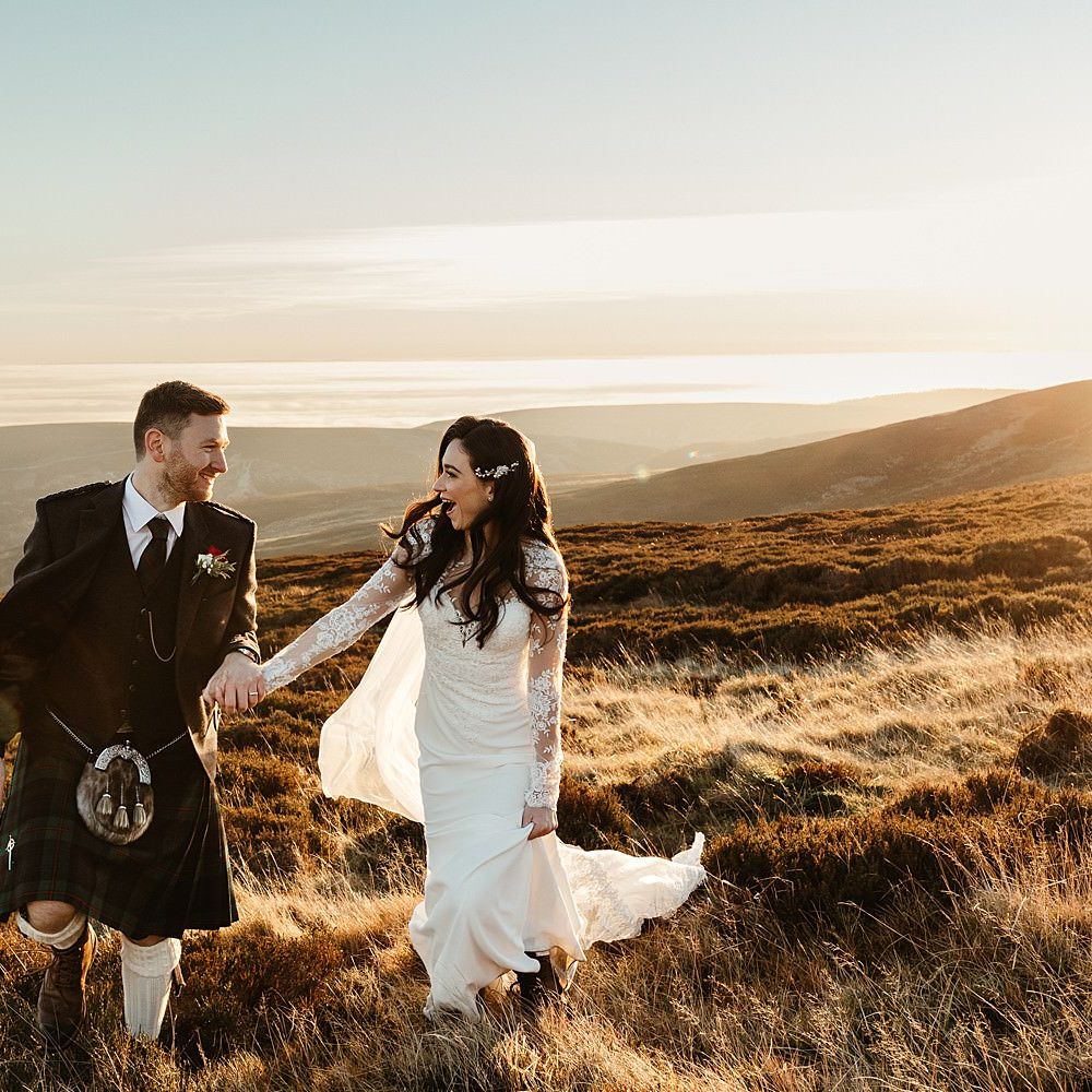 Bride & groom walk across hillside in front of beautiful Scottish highlands whilst the sun sets 
