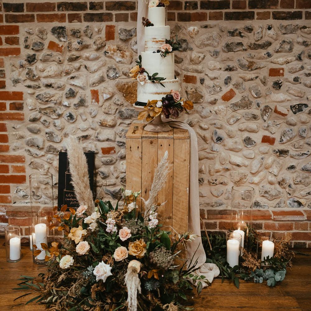Four tier white frosted rustic wedding cake with autumnal flowers and a personalised cake topper on a wooden crate with candles 