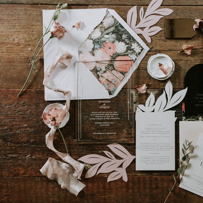 flat lay of romantic wedding stationery suite with floral envelopes, acrylic invitation and pink ribbon
