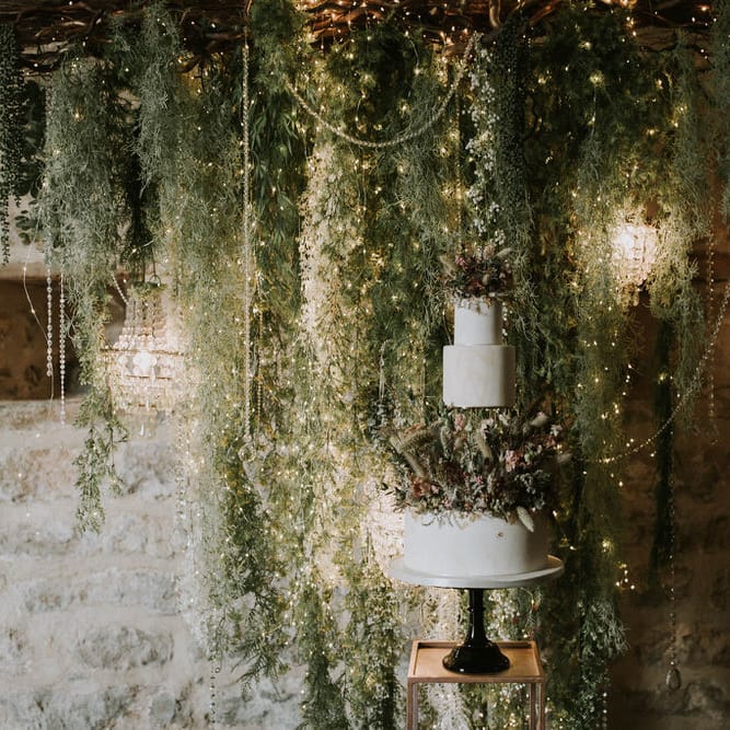 three tier wedding cake decorated with dried and fresh flowers in front of a rustic twig, foliage and fairy light backdrop 