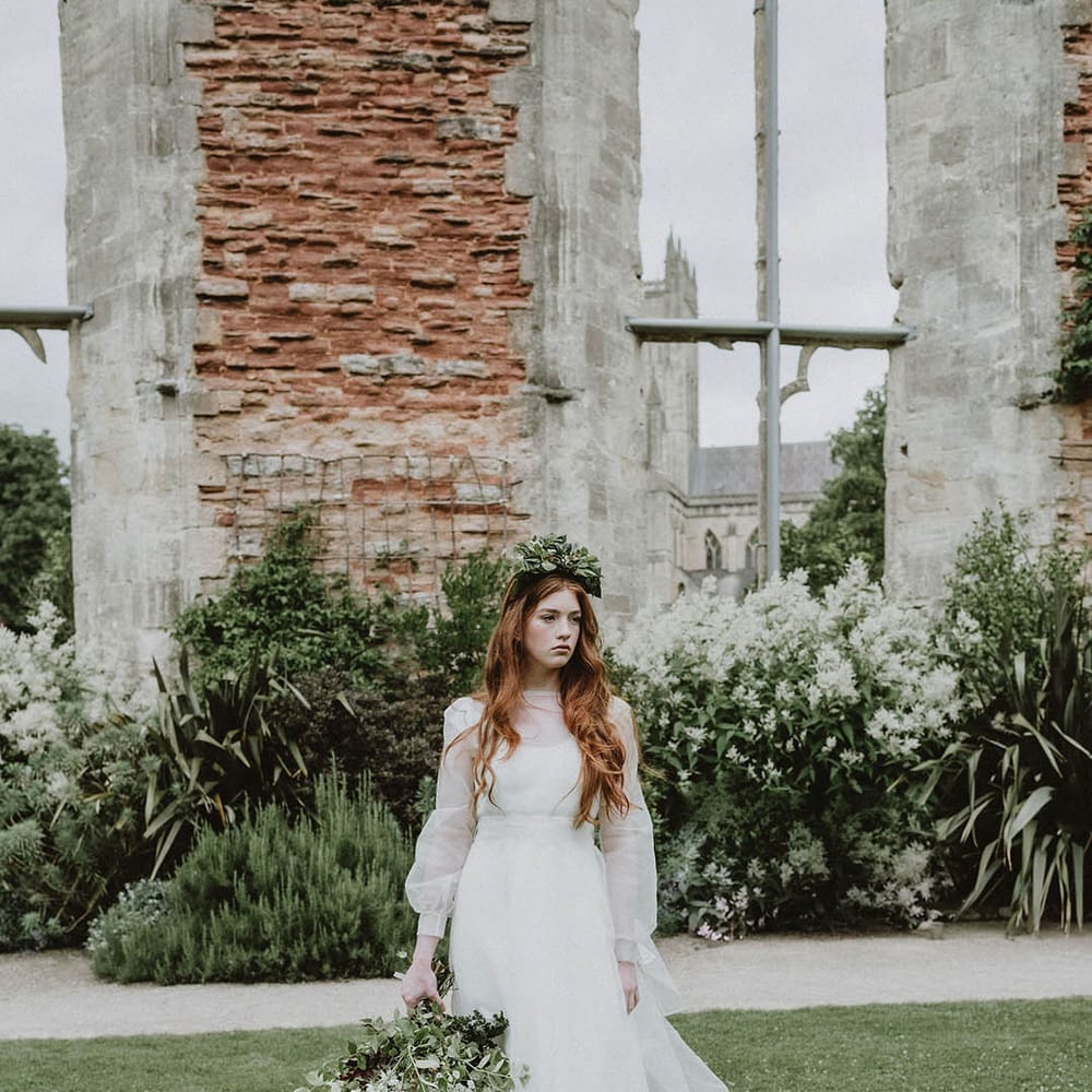 Bride in a wedding dress with sheer layer wearing a foliage flower crown standing in the gardens at Bishop's Palace, Wells