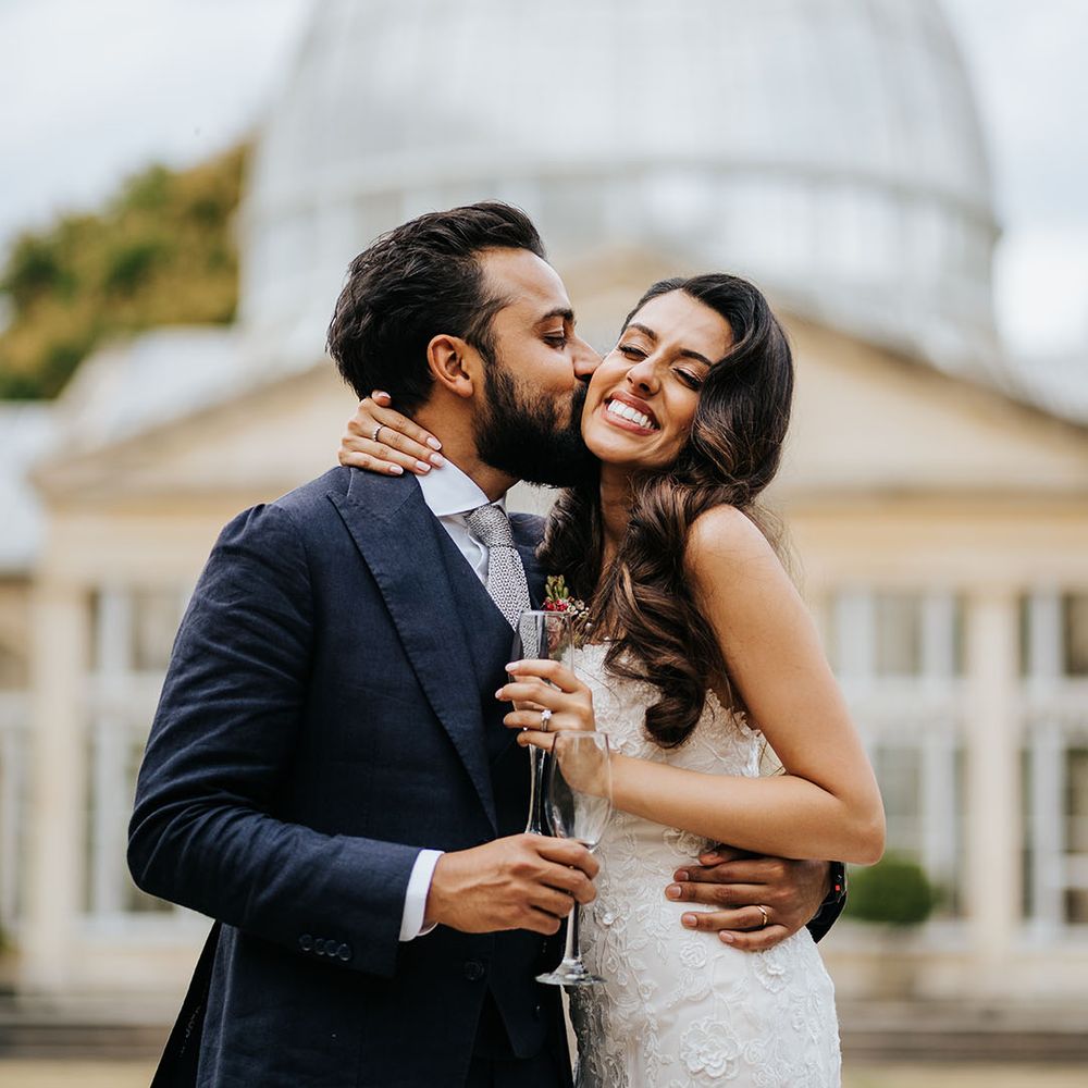 Bride & groom hold champagne glasses as groom kisses his brides cheek on the day of their wedding