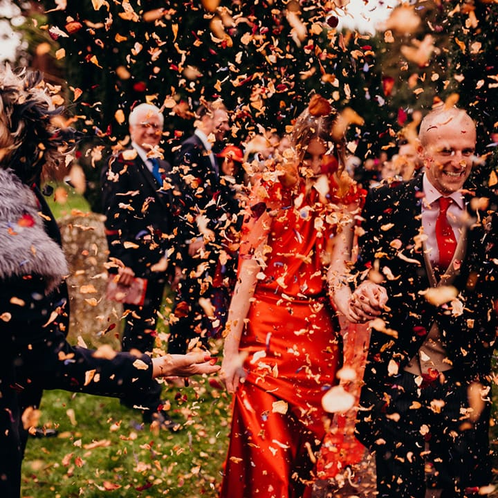 Bride in red satin wedding dress with cape and groom in three piece suit with red tie holding hands doing the confetti walk at winter wedding