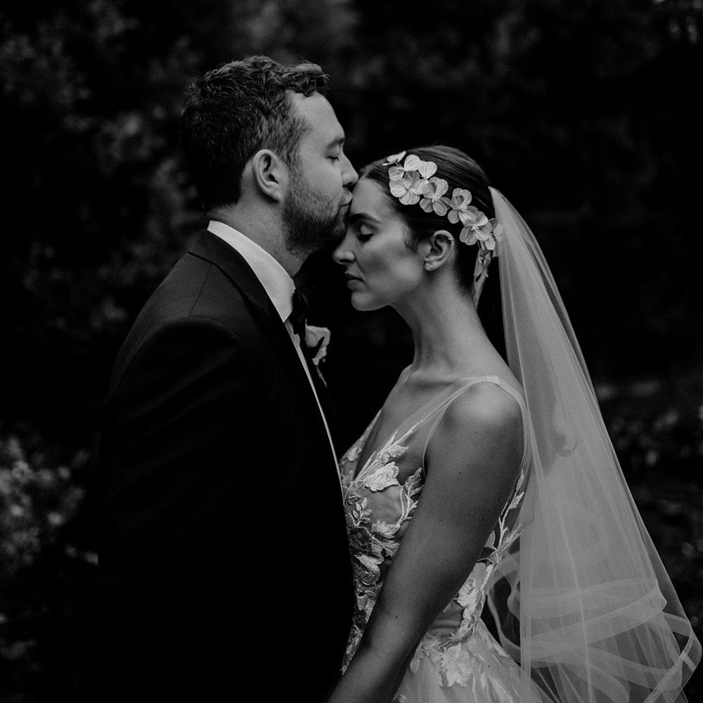 Groom in black tie kisses the bride on the forehead wearing a flower wedding dress 