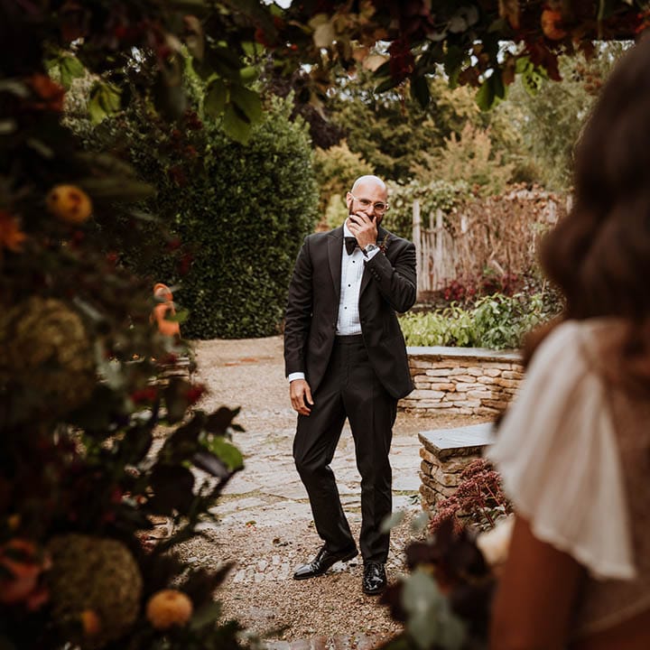 First look moment; Groom in classic black tuxedo with black bowtie looking at bride in open back detail lace wedding dress