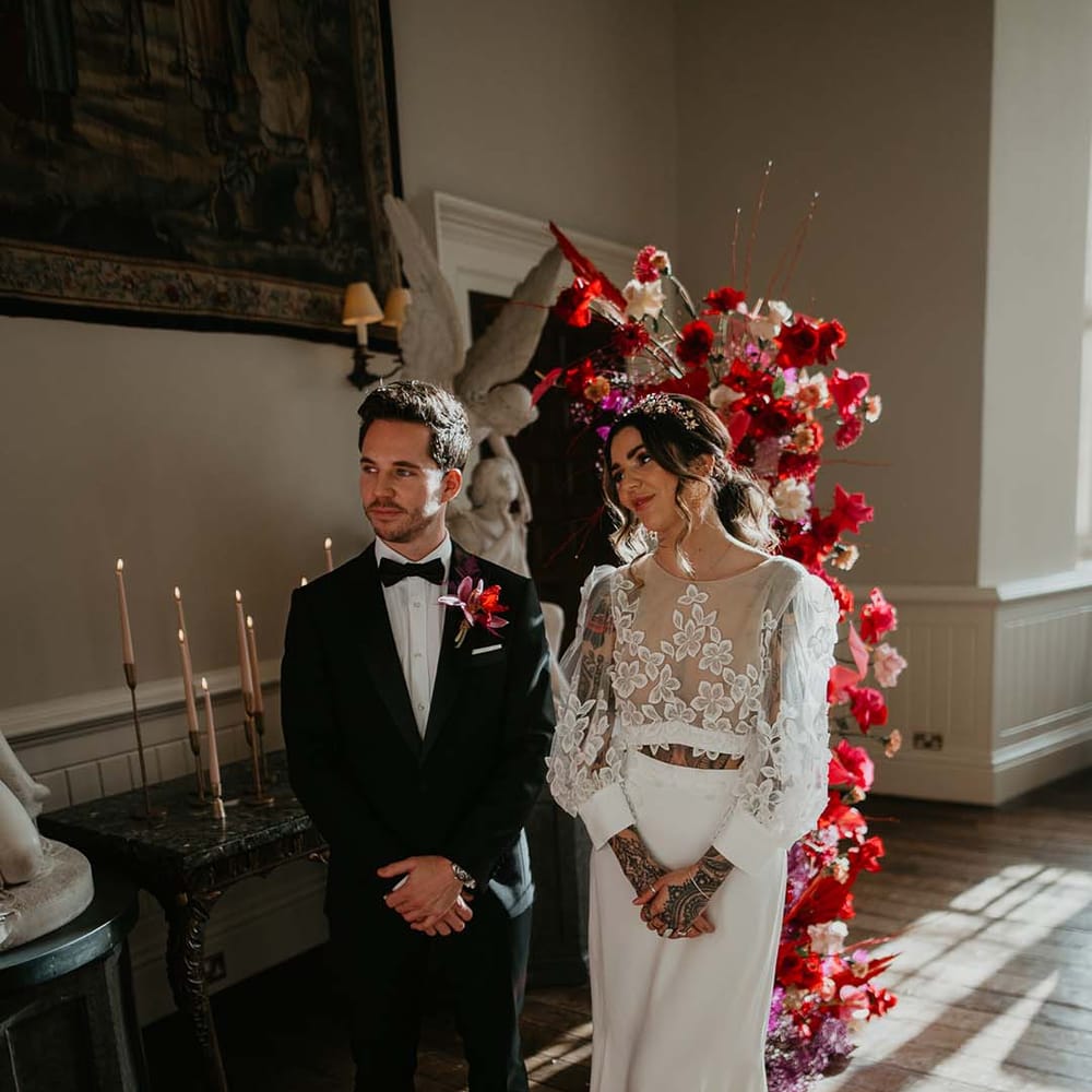 Bride in two piece bridal separates with puff sleeve sheer crop top with 3d appliqué flowers and full bodied silk skirt and groom in black tuxedo with magenta amaryllis and orchid boutonniere standing at the alter of Elmore Court