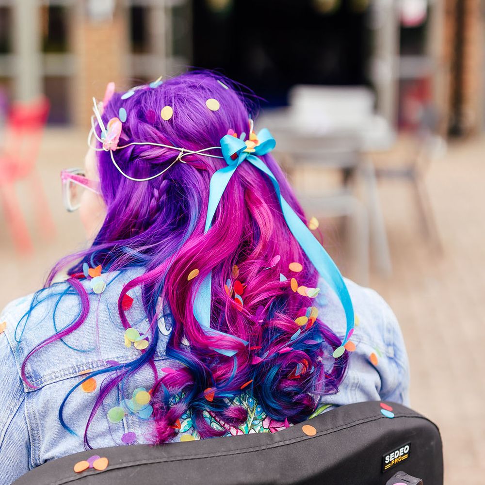 Bride with vibrant purple hair with blue bridal hair ribbon and confetti in wheelchair with blue glittery fairy wings 