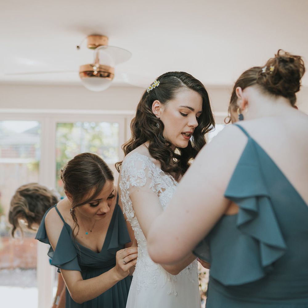 The bride gets ready for the wedding day with daisies in her hair and the bridesmaids helping her into her beaded wedding dress 