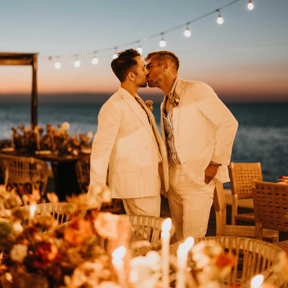 Groom in mens white wedding suit kissing groom in matching suit under suspended fairy light wedding lighting by rustic burnt orange wedding tablescapes 