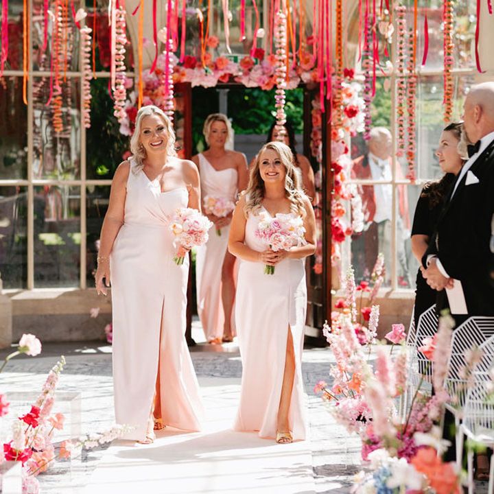 Bridesmaids walk down the aisle in pairs in one shoulder pink bridesmaid dresses 