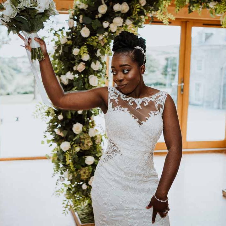Bride entering wedding breakfast wearing lace and mesh boatneck wedding dress with silver bridal hair accessories holding large white rose and foliage bridal bouquet 