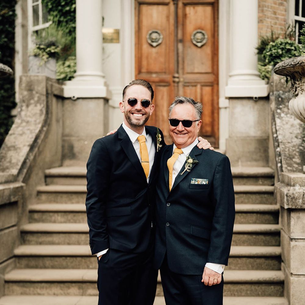 Groom and father of the groom in matching navy suits with yellow ties wearing sunglasses 