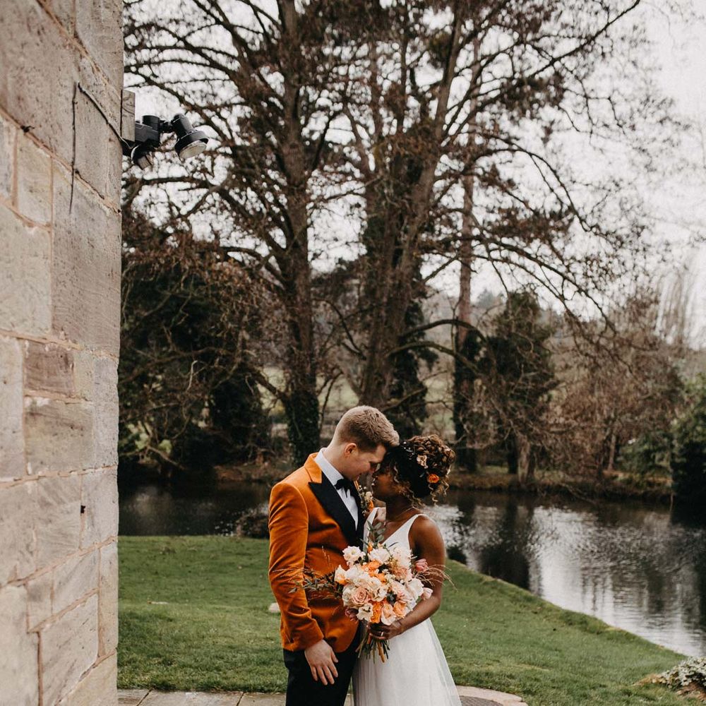 Groom in crushed burnt orange velvet grooms blazer, black bowtie and black suit trousers embracing bride in v neck sleeveless wedding dress with 3D floral applique train holding blush and orange garden roses, poppies and carnations, dried flowers and eucalyptus wedding bouquet at Brinsop Court wedding venue