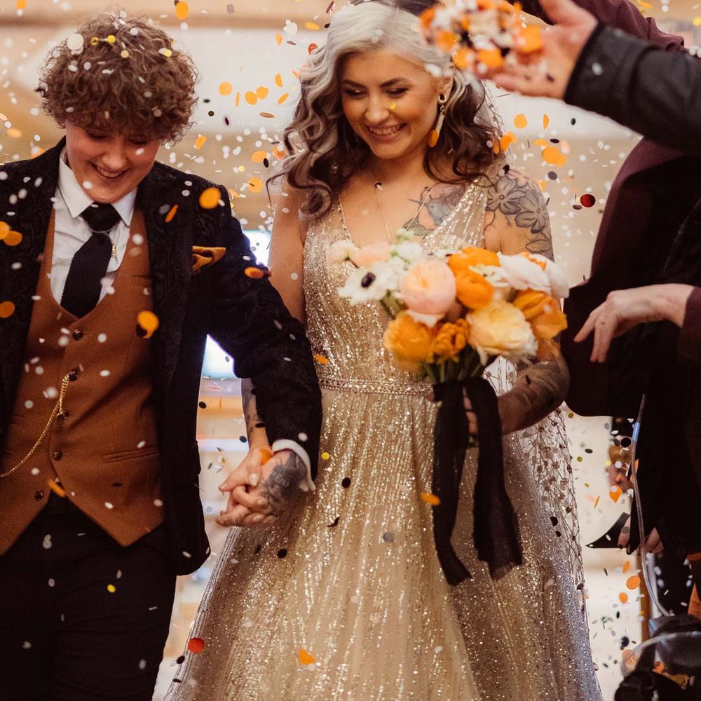 Bride in sleeveless deep v neck sparkly gold wedding dress with sheer and gold polka dot wedding veil holding orange garden rose, white poppy, light pink carnation and foliage bridal bouquet wrapped in sparkly gold ribbon doing colourful wedding confetti exit with bride in dark velvet bridal jacket with rust waistcoat and pocket square, black suit trousers and gold pocket watch 