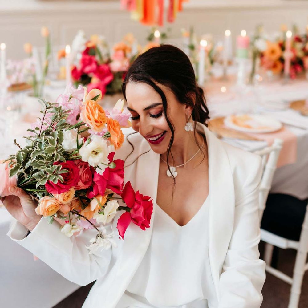 Bride in white bridal suit with silver bridal jewellery holding mixed seasonal bridal bouquet sitting in by wedding tablescape with white wedding tablecloth, burlap place mats, tapered candles, pink and orange wedding stationery, and floral wedding table centrepieces with pink and orange garden roses, orange floribunda, yellow ranunculus flowers, foliage and Iceland poppies with red, pink orange and yellow balloons and wedding streamers at Merriscourt wedding venue