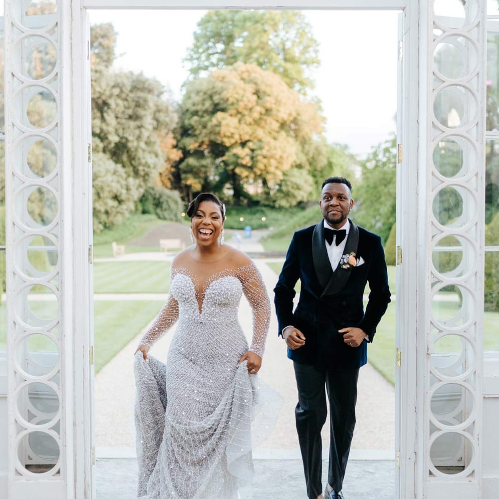 Groom in velvet midnight blue double breasted groom’s blazer, velvet bowtie and peach garden rose boutonniere walking into Syon Park reception room with bride in illusion v neck sheer off shoulder long sleeve pearl embellished wedding gown 