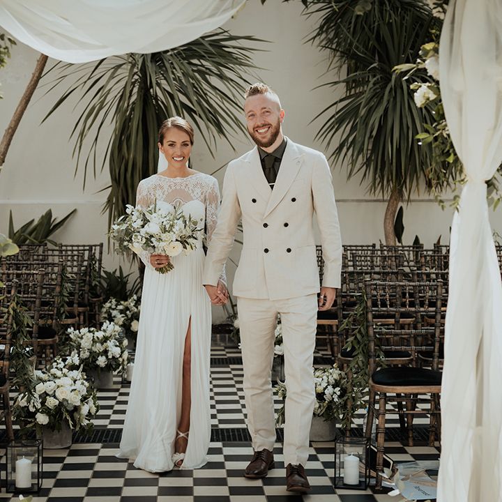 groom in a double breasted beige wedding suit holding hands with his bride in a. Catherine Deane wedding dress under a floral drape altar at Gunnersbury Park 