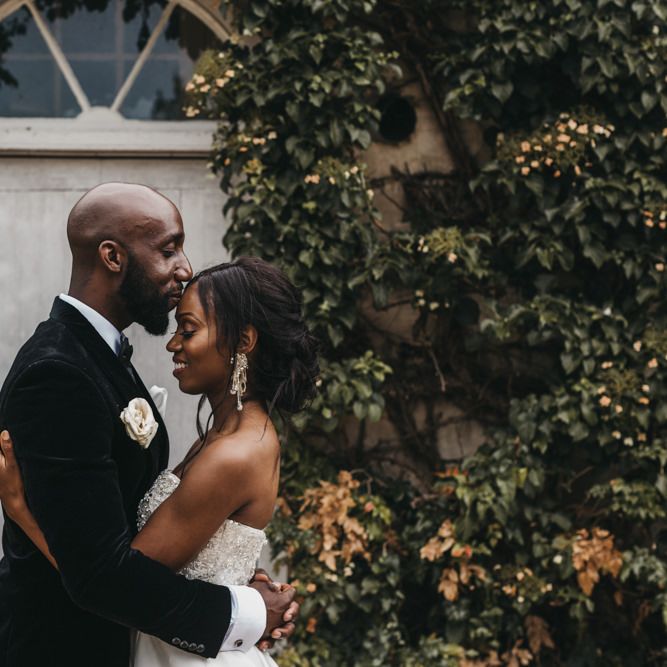 Black bearded groom in a velvet tuxedo kissing his brides forehead in a strapless Oleg Cassini wedding dress 