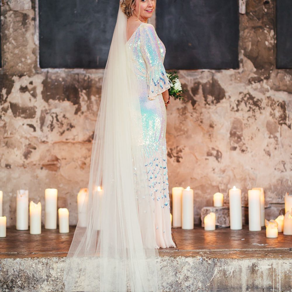 Blonde bride wearing iridescent dress looks back at camera surrounded candles