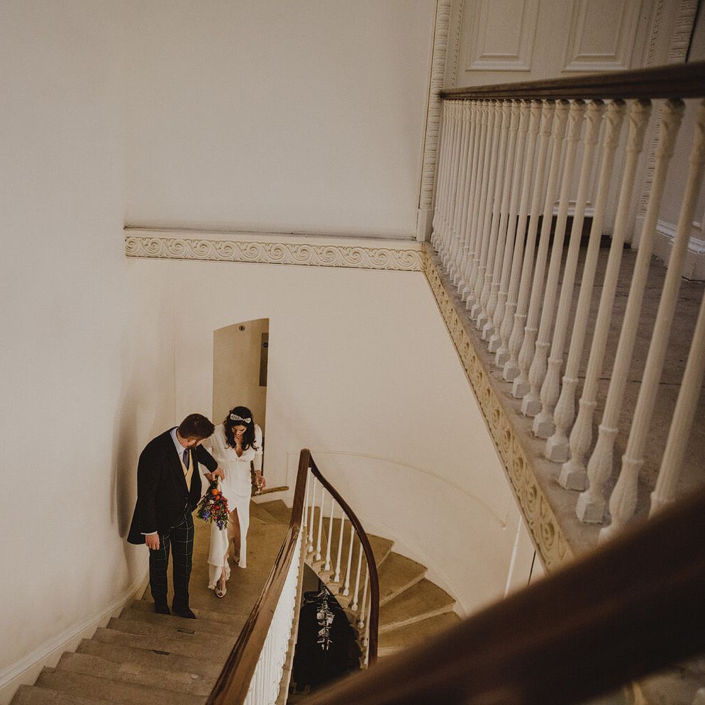 Bride and groom walking up stair case at London wedding bride wears long sleeve front slit wedding gown holding bright flowers, groom wears tartan trousers 