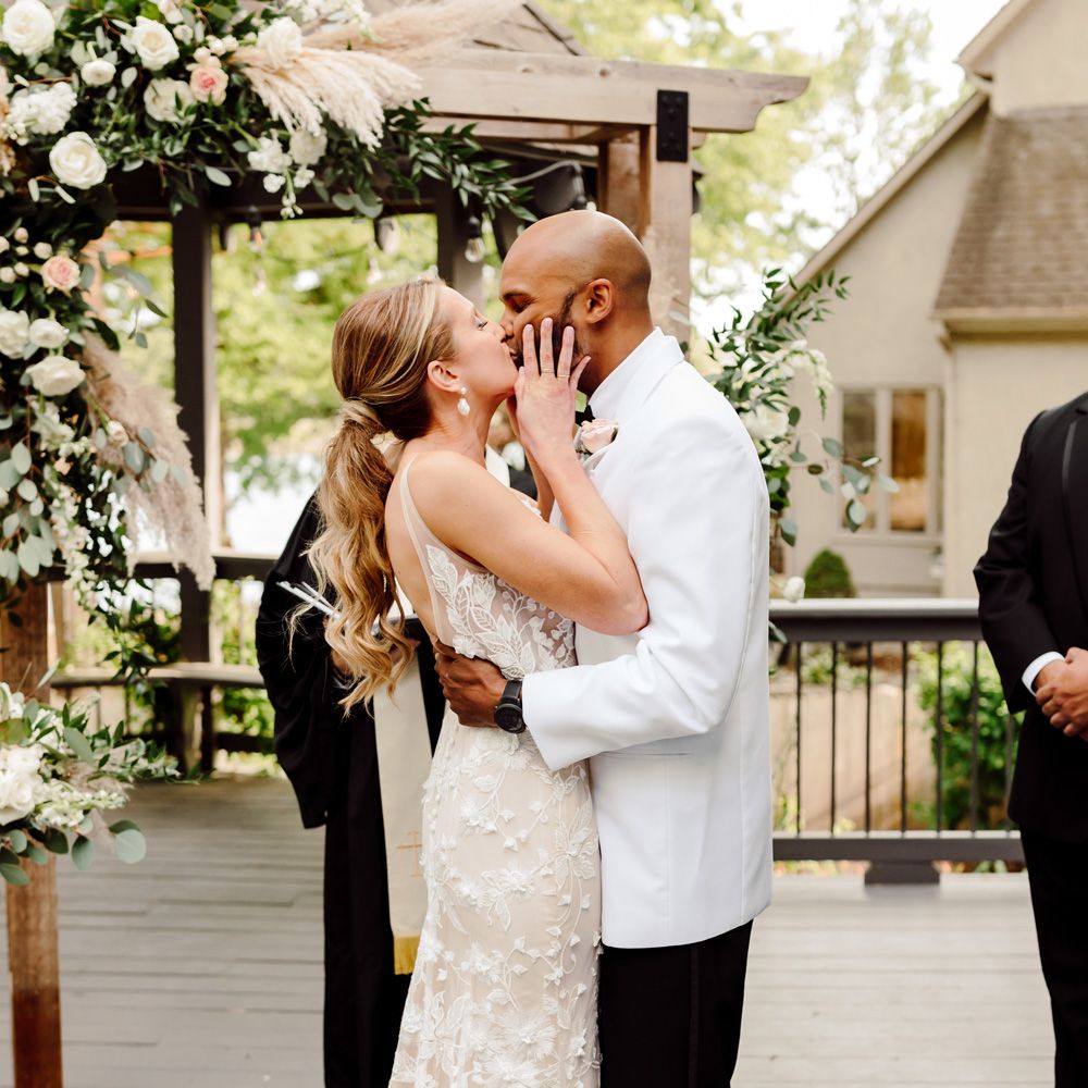 Bride in white embroidered Made With Love wedding dress and curled ponytail embraces groom in white tuxedo jacket during wedding ceremony at backyard wedding