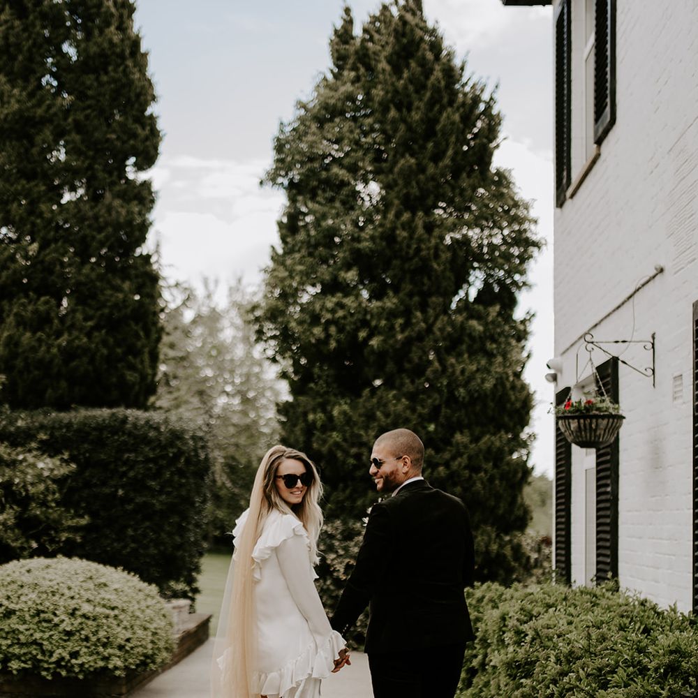 Stylish bride in a short wedding dress with long dusky pink veil and sunglass walking through the grounds of Waresley Park Estate with her groom 