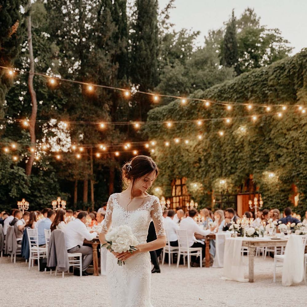 Bride stands outdoors in Italy wearing lace wedding gown and fairy light canopy  | Royal Studio