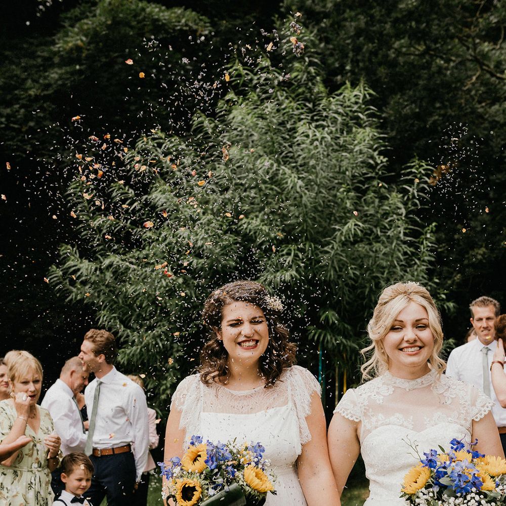 Smiling brides walk through confetti together as a married couple with sunflower, gypsophila and bellflower bouquets