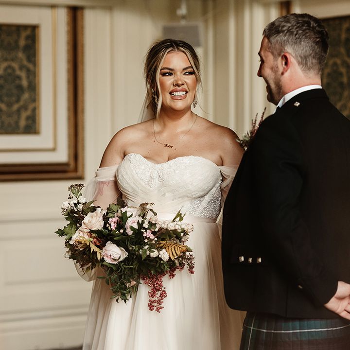 Bride and groom smile at each other with bride in custom wedding gown with sheer sleeves and autumnal classic wedding bouquet 